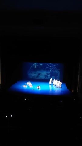 A video displaying a stage performance in a dark theater. The stage is lit with vibrant blue and white lights. In the center of the stage, a projection displays a deep blue underwater scene with what appears to be a large moon or planet. Several figures, possibly dancers or performers, dressed in white, are positioned in front of the projection. Some of these figures are partially obscured by the lighting. The overall scene is illuminated by the stage lights, creating a dramatic and somewhat ethereal atmosphere.