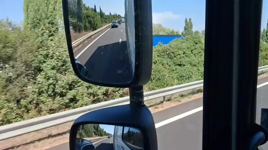 The video shows a view from inside a vehicle, looking forward. The vehicle is on a highway, with a clear blue sky and cypress trees lining the road. There are other vehicles visible: a green car is in the left lane, and a white truck with "VOULAND FERS" written on the back is in the right lane. Another car is behind the green car. The green car appears to be merging into the right lane, in front of the truck. The camera is positioned to capture the road ahead and the surrounding scenery.