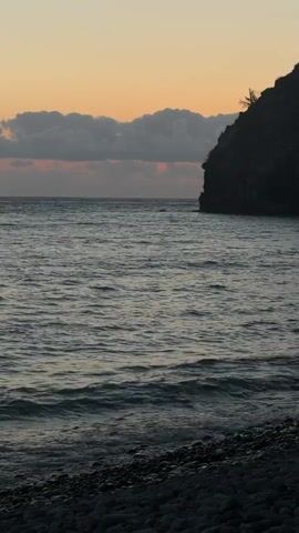 The video shows a tranquil ocean scene at dusk. The sky is a warm gradient of orange and yellow, with soft, gray clouds drifting across the horizon. In the foreground, gentle waves lap against a pebble beach, creating a soothing rhythm. To the right, a dark, silhouetted cliff face rises from the water, its steep incline softened by the dimming light. A single palm tree is visible at the very top of the cliff. The water itself is a deep, calm blue, reflecting the muted colors of the sky. The overall impression is one of peace and natural beauty.