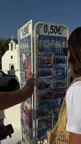 Here's a detailed description of the video:

The video shows a person looking at postcards on a rotating display rack. The rack is white with metal shelves, and a sign on top displays the price of 0.50€. The postcards show various scenes, including landscapes, seascapes, and town views.

One person, likely a customer, reaches out and touches several of the postcards. Beside that person stands another person with their back to the camera, wearing a white shirt and a woven straw bag. 

In the background, there is a white building with a red roof, possibly a church, and a dark-colored car. The sky is clear and blue, suggesting a sunny day. The environment appears to be an outdoor tourist area, possibly in Greece.
