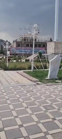 This video captures a view of a public square or monument area in Ismoili Somoni, Tajikistan. The primary subject is a tall, slender monument topped with a dome, featuring a statue of a standing figure at its base. The monument is set against a backdrop of buildings and a mural or series of artworks depicting scenes and figures. The foreground is dominated by a large, paved area with a geometric tile pattern.

The scene appears to be set during the daytime, under a cloudy or overcast sky. There are lampposts and decorative white fencing along the sides of the paved area, suggesting a public park or plaza designed for gathering. Greenery and some rose bushes are visible around the monument and to the sides, indicating a maintained outdoor space. There are no people clearly visible in the immediate frame, though the setting suggests it's a place where people might gather.

The overall atmosphere seems calm and perhaps a bit solemn, given the monumental nature of the central structure. The location cues, such as the architectural style and the specific monument, point to a historical or cultural site within Tajikistan. There is some text visible on banners or signs in the background, but it is too small and blurry to be deciphered.