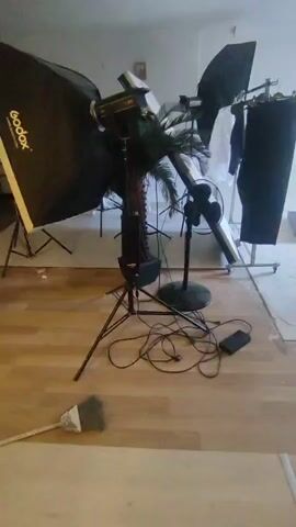 The video pans across a room that appears to be set up for a photoshoot. In the foreground, a broom lies on the wooden floor. Dominating the center of the frame is a photography lighting setup, including a large softbox on a tripod, a second light with a smaller modifier, a palm tree in a pot, and a black standing fan. Cables are strewn across the floor, connecting the equipment. In the background, a clothing rack holds several garments, and a small framed picture hangs on the wall. The room seems to be in a state of preparation or disarray, with some parts of the floor appearing unfinished.