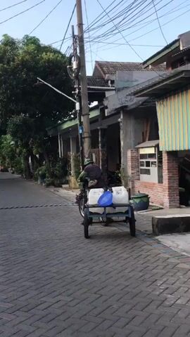 A person on a motorcycle is pulling a trailer with two large white containers and a blue lid. The motorcycle and trailer are on a paved street lined with houses and trees. The person is wearing a hat and a dark shirt. The motorcycle is moving forward. The sun is shining.
