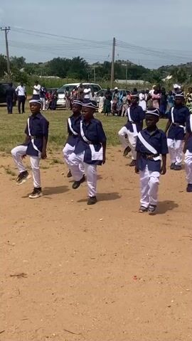A group of eight young boys, all wearing matching navy blue uniforms with white sashes and white hats, stand in formation on a dusty, outdoor field. They are facing the camera and appear to be participating in a parade or ceremonial event. The boys are of similar age and build. Behind them, a crowd of onlookers, some sitting and some standing, are visible. Several vehicles, including a white van and a car, are parked in the background. The environment appears to be a rural or semi-rural area with some vegetation and a few buildings or structures in the distance. The sky is overcast with some hints of blue. The boys are standing at attention with their hands at their sides, and their expressions are neutral.