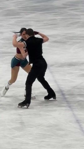On an ice rink, a pair of figure skaters are performing a lift. The female skater is wearing a two-piece skating costume with burgundy and green fabric, and knee-high socks. The male skater is wearing a black vest over a white shirt, and black pants. During the lift, the male skater holds the female skater up, her body upside down and outstretched. After the lift, they both turn and skate away.