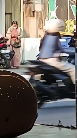 A street scene with several people and vehicles. In the foreground, a brown helmet with white dots is partially visible. In the mid-ground, a person on a blue motorcycle is seen from the side, wearing a dark jacket, white pants, and a hijab. Next to them, a white SUV is parked, with its rear visible. Further back, a person in a light-colored jacket and pink pants stands near a pile of cardboard boxes. A blue trash bin is visible on the right. In the background, a white truck with red taillights is driving away. Several other motorcycles are parked behind the blue motorcycle. The overall environment appears to be an outdoor urban setting with buildings in the background. The video has a slight blur due to movement.