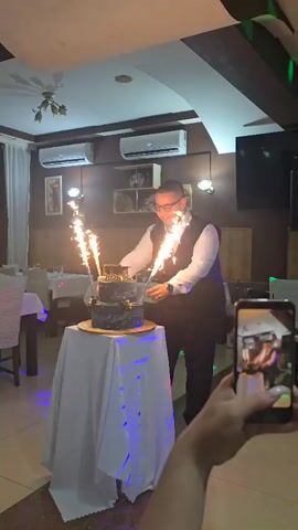 A man in a white shirt and black vest smiles and poses with four sparklers, two in his hands and two on a three-tiered cake, as a person in the foreground records him with a phone. The man raises his arms with the sparklers and glances to his right, while the cake on a white table cloth has blue and purple lights projected onto it. The background shows an empty restaurant with tables and chairs.