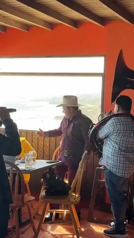 A man wearing a white cowboy hat and a maroon patterned shirt with burgundy pants is standing near a table and a man playing a tuba. The man in the cowboy hat is looking out a large window at a body of water. A woman wearing a yellow shirt is seated at the table. The room has orange walls and a wooden ceiling. There is a large tuba instrument on the right side of the frame.