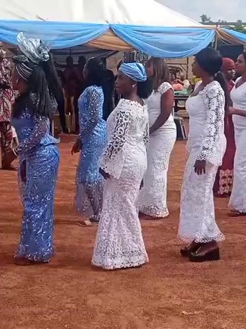 A medium shot captures a group of men and women dressed in white and blue attire gathered for a celebration. A man in a white kaftan and black cap walks through the frame, followed by a man in a white kaftan with a patterned blue and green headpiece. Another man in a white kaftan walks past the camera, and then a woman in a long white dress. The ground is reddish brown, and a white tent with blue and brown drapes is visible in the background. A metal frame decorated with white flowers and green leaves stands to the right.