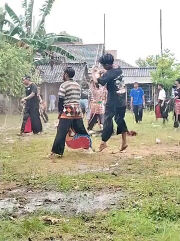 The video shows a group of men in a grassy, muddy area. Some are wearing black pants and tops with red wraps around their waists. One man is wearing a horizontally-striped shirt and black pants with a more elaborate waist wrap. Another is wearing a blue shirt and black pants. They are barefoot and appear to be performing a dance or martial arts routine, with movements involving jumping, kicking, and whipping the ground. The setting appears to be outdoors, in a yard, with a building in the background. It appears to be raining. There is also a person partially visible in the foreground, holding what looks like a drum with the word “KENANGAN” on it.