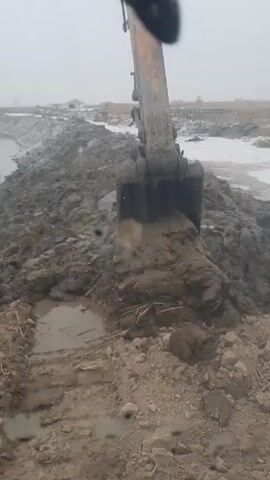 An excavator is digging into a bank of wet mud. The bank is next to a narrow waterway. The sky is overcast and a light snow or rain is falling. There is a house or shed in the distance across the waterway.
