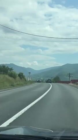 The video captures a first-person perspective from a vehicle traveling on a two-lane asphalt road through a mountainous landscape in Plugova, Romania. The road features a solid white line down the center, indicating a no-passing zone. Ahead, several cars, predominantly dark-colored SUVs, are visible, along with a white truck on the left side of the road in the distance.

Lush green mountains and hills rise on both sides of the road, extending into the background. The sky is partly cloudy, with patches of blue visible between white clouds, suggesting a bright daytime. Utility poles and power lines are present along the roadside. A hint of a red barrier or fence can be seen on the right side of the road towards the end of the clip. The road appears to curve gently to the left as it traverses the scenic terrain.
