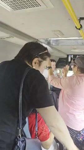 The video is shot from inside a bus, looking forward and slightly to the right. The camera is positioned behind a person wearing a black t-shirt, who is facing away from the camera. This person's left arm is resting on the back of a seat. In front of them, to the right, another person wearing a pink shirt and a grey baseball cap is visible, facing left and extending their right arm forward, with their hand raised as if to block something or make a gesture. Another person, partially obscured, is behind the person in the pink shirt. Further ahead, a person with blonde hair wearing sunglasses is seated on the right side of the bus, looking to their left. The bus interior shows yellow handrails, air vents, and grey seats with blue headrests. The lighting is dim, suggesting it might be daytime but with the bus interior being relatively dark.