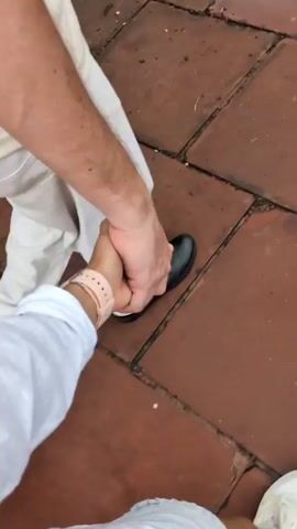 The video shows a close-up perspective of two people holding hands and walking on a terracotta tiled surface. The person in the foreground, from whose perspective the video is filmed, is wearing a white shirt and a light pink watch on their left wrist. Their hand is clasped with the hand of the person in the background. This second person is wearing white pants and black shoes. The camera angle is low, focusing on the legs and hands of the individuals. The terracotta tiles appear to be slightly wet, suggesting recent rain or a damp environment. The overall scene depicts a casual, possibly romantic, interaction between two people walking together.