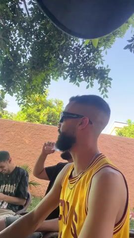 The video shows a man with a beard and sunglasses, wearing a yellow basketball jersey and a silver chain, looking at the camera and laughing. He is sitting in what appears to be an outdoor patio or backyard, with trees providing shade. Other people are sitting nearby, one holding a water bottle, and there is a low peach-colored wall in the background. The sky is blue.