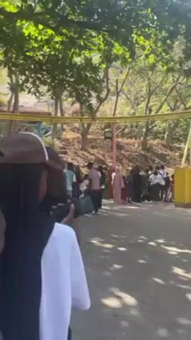 The video shows a crowd of people gathered near a fairground ride. The ride structure is visible, painted in bright colors like yellow, purple, and blue. Individuals of various ages and clothing styles are present, including some wearing hijabs. Some people appear to be queuing or waiting near the ride, while others seem to be walking around. The background features trees and a hillside. A uniformed worker is visible in the middle of the frame.  Food stalls are visible under the ride structure.
