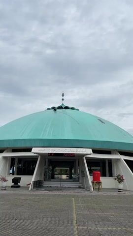 A person is seen on the turquoise dome of a mosque, performing some task. Another person is visible further up the dome near the apex. The mosque is identified by text above the entrance as "MASJID RAUDHATURRAHMAN". Below the mosque name, a digital display reads "Selamat Datang di Masjid Besar". The sky is overcast. In front of the mosque entrance, there are stairs, railings, and a red water tank on a stand.