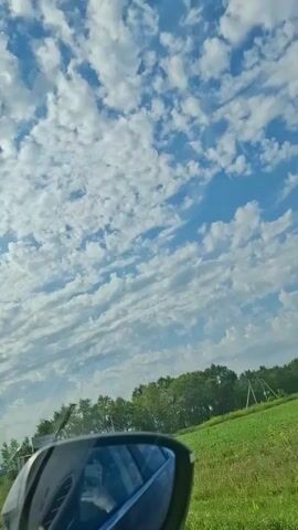 The video is shot from inside a vehicle, looking up at the sky. The sky is blue with many fluffy white clouds. On the right side of the frame, there is a green field and trees. A side view mirror is visible in the foreground. The vehicle appears to be moving.