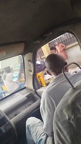 The video is taken from inside a vehicle, looking out through the windshield and side window. In the foreground, the back of a person's head and shoulders are visible, wearing a light-colored shirt. The interior of the vehicle appears worn. Through the window, a street scene unfolds. To the left, a person in a purple shirt is looking towards the viewer. Further to the right, a person in a peach-colored outfit is standing near a fence. In the middle ground, a street vendor's stall is visible, displaying fruits such as pineapples and watermelons. A bicycle with a yellow jerrycan and a black bucket strapped to its rack is parked nearby. A woman in a white t-shirt is interacting with the vendor. The environment suggests an outdoor market or busy street in a town or city. The lighting indicates it is daytime.