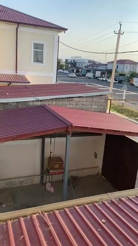 A video shows a street view from what appears to be a second-story window or balcony. In the foreground, a burgundy metal roof is visible. Below that is another structure with a burgundy metal roof and walls, featuring a doorway on the right side and an open space on the left with a table and a suspended object. Beyond this, across a road, there are several houses with burgundy roofs and a gas station on the right. Various vehicles, including cars and vans, are seen on the road and parked. Telephone poles with wires stretch across the scene. The sky is clear and appears to be either early morning or late afternoon.