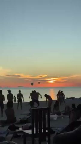 The video shows a group of people playing on a beach at sunset. The sun is setting over the ocean, creating a warm, orange glow. There are silhouettes of people playing with a ball on the beach, and others relaxing in the foreground. A few chairs and beach bags are visible. The sky is a clear blue, transitioning to orange near the horizon.