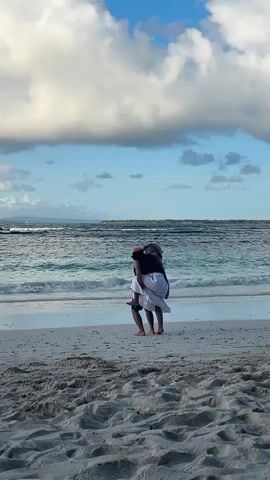 Two people are standing on a sandy beach with the ocean in the background. The person in the front is wearing a hat, sunglasses, and a vest over a shirt and shorts, and appears to be dancing. The person behind them is wearing a hat and a white skirt and is also facing the water. The sky is blue with scattered clouds.