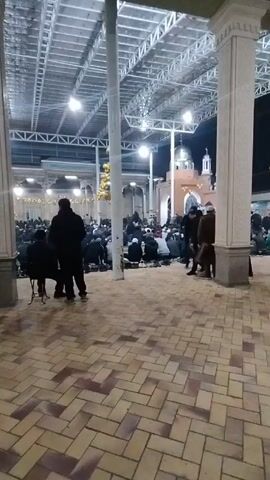 The video shows a gathering of people inside a large, open structure. Many individuals are kneeling on the ground, seemingly in prayer or meditation. Others are standing in small groups, talking and interacting with each other. The structure is supported by large columns and has a high, ornate ceiling with visible metal framework. The floor is covered in a patterned tile design of light and dark brown. The overall atmosphere appears to be one of reverence and community.