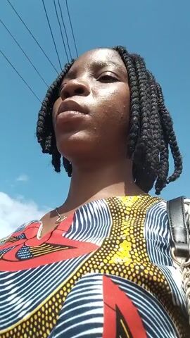 The video is a selfie-style shot of a young black woman with braided hair and a colorful African print dress. The camera is angled upwards, showing her face and upper body against a bright blue sky with some power lines visible. She appears to be looking upwards and slightly to the side, with a neutral or slightly thoughtful expression. The lighting suggests it is daytime.