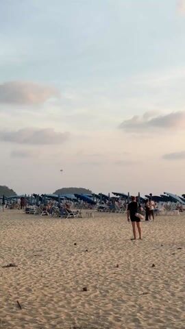 The video shows a beach scene. Initially, the beach is populated with people, beach chairs, and umbrellas. Two people walk across the sand in the foreground. The camera pans to the right, showing people walking along the shore, and eventually focuses on a group of people playing volleyball. The sun is setting in the background, creating a golden hue.