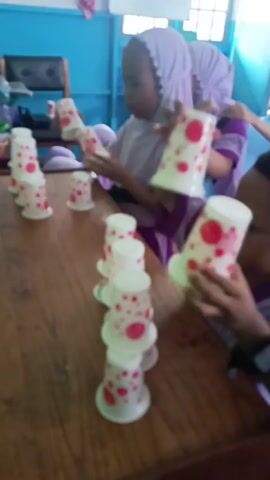The video shows three young girls in a room, likely a classroom, with blue walls and a wooden table. The girls are wearing hijabs and appear to be engaged in a cup-stacking activity. There are many white plastic cups with red polka dots stacked in towers on the table. The girl in the foreground on the left is wearing a purple hijab and a purple and white shirt. She is holding a cup and looking at the tower she is building. The girl in the middle is wearing a light purple hijab and a purple shirt. She is looking at the cups and seems to be in the process of stacking them. The girl on the right is wearing a light purple hijab and a purple shirt. She is holding a cup and looking towards the camera. The table is made of wood and has a worn appearance. The lighting in the room is natural, coming from a window that is not visible in the frame.