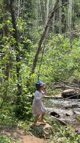 A young child wearing a light-colored dress and a blue hat stands on a rock near a stream in a forest. The child is facing right and appears to be dancing or playing. The forest is lush with green trees and foliage, and there are rocks and fallen branches around the stream. The sun is shining through the trees.