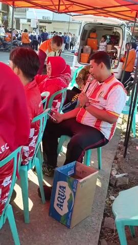 A man in a white, red, and yellow striped shirt is sitting in a teal plastic chair and speaking into a microphone. He is holding the microphone in his right hand. He is looking to his left. To his left, a woman in a red hijab and a red jacket is sitting in a teal plastic chair, looking at her phone, which she is holding in her left hand. Behind the man, a white van is parked. Boxes are visible in the back of the van. Several people are standing around the van and in the background. A red tent is erected over the scene. A cardboard box with an Aqua logo is on the ground next to the man. The scene appears to be outdoors during the day.