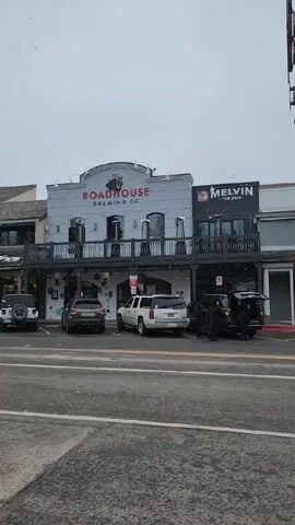 The video shows the street scene in front of Roadhouse Brewing Co. and other shops on a snowy day. Cars are parked along the street in front of the businesses. There are two people near the trunk of an SUV in front of Melvin. The camera pans left to show the other storefronts on the street. Snowflakes are visible.