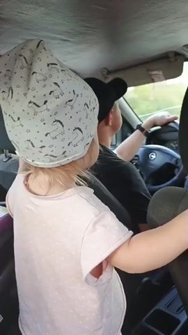 A young child wearing a light pink t-shirt and a white hat with a unicorn pattern stands in the back seat of a car. The child's hands are resting on the back of the driver's seat. The driver, wearing a black baseball cap and a dark shirt, is visible from the shoulders up, gripping the steering wheel and looking forward. The car interior appears to be a typical passenger vehicle, with visible seat upholstery and dashboard elements. The lighting suggests it is daytime, with some light filtering in from outside. The video is captured from a low angle, focusing on the child and the driver's interaction.