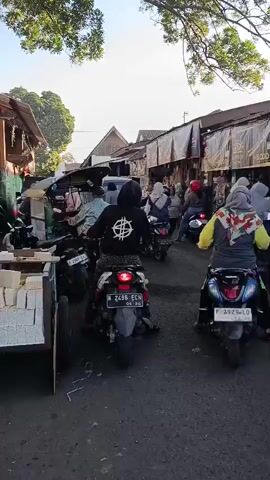 The video shows a street scene with several motorcycles and people. The camera is positioned behind a person on a motorcycle, facing forward. In the foreground, there is a motorcycle with a license plate that reads "N 2496 ECH". The rider of this motorcycle is wearing a black hooded sweatshirt with a white circular symbol on the back. To the left of this motorcycle, there is a cart with white objects, possibly food items. Behind the lead motorcycle, other motorcycles and people are visible. Some people are walking, while others are on motorcycles. The street is lined with buildings that appear to be shops or market stalls. The sky is visible at the top of the frame, with some trees and branches extending into the scene. The overall environment suggests a busy street, possibly a market, with moderate traffic. The lighting indicates it is daytime, possibly morning or late afternoon due to the angle of the sun.