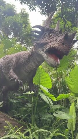 A close-up, low-angle shot shows a realistic dinosaur replica with brown, scaled skin and prominent horns on its head. The dinosaur's mouth is open, revealing a red interior, and its eyes are dark. It is positioned amidst lush green foliage, including large-leafed plants and ferns, with trees and a bright sky visible in the background. The lighting suggests an outdoor environment, possibly a park or exhibit, during daylight.