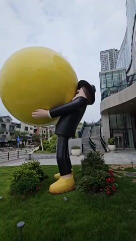 A full shot of a large, yellow, spherical object being held by a cartoonish figure with a black suit, black hat, and yellow shoes. The figure is bent over, appearing to struggle with the weight of the sphere. The scene is outdoors, with a sidewalk, grass, bushes, and a street in the background. Buildings and a cloudy sky are also visible. The video pans slightly to the right.