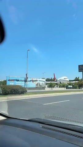 The video is taken from inside a vehicle, looking out through the windshield. A bright blue sky dominates the upper portion of the frame. In the distance, a large white airplane with a red tail fin is visible, positioned behind some trees and buildings. The airplane appears to be stationary, possibly on the ground at an airport or as a static display. On the left side of the road, there are advertisements and a tall light pole. On the right side of the road, there are more trees and some cars driving by. The road itself is paved with lane markings visible. The overall environment suggests a sunny day in an urban or suburban area, likely near an airport or transportation hub.
