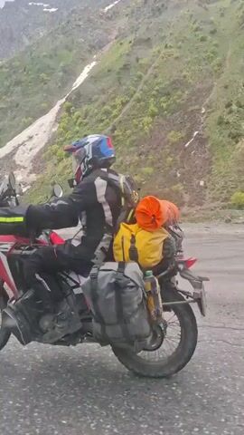 The camera is mounted on a moving vehicle, possibly a motorcycle, as indicated by the presence of a red fender and tire visible on the right side of the frame. The vehicle is moving along an asphalt road. In the background is a mountainous landscape. The lower slopes of the mountain are covered in green vegetation. There are patches of snow visible along the mountain side. The peak of the mountain is obscured by cloud cover, but appears to be rocky and potentially snow-covered. The weather seems fair, with patches of blue sky visible between clouds.