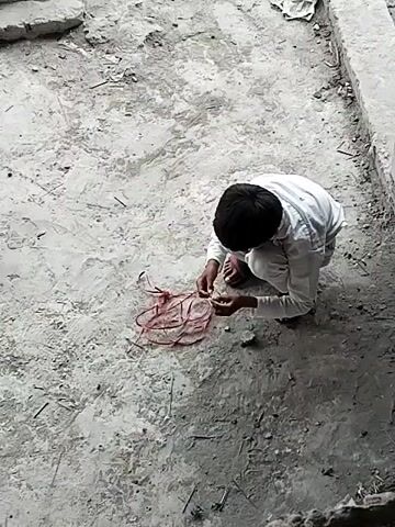 The video shows a young boy, likely a child, crouched on a dusty, grey concrete surface in an outdoor setting. He is wearing a light-colored, long-sleeved shirt and appears to have dark hair. His back is mostly facing the viewer, but his head is tilted down, indicating focus on an activity.

In front of him, on the ground, is a tangled mass of red string or thin wire. The boy is using both hands to manipulate this material, seemingly trying to untangle it, tie a knot, or play with it. His posture suggests deep concentration on the task.

The immediate surroundings feature a rough, utilitarian concrete ground, with some debris and specks visible. In the background, there are hints of concrete structures, possibly low walls or foundations, and other indistinct materials suggesting a modest or undeveloped outdoor area. The lighting indicates it is daytime, though no direct sunlight or strong shadows are present, suggesting an overcast day or an area partially shaded. There is no visible text. The scene conveys a sense of a child finding engagement in simple play within his environment, consistent with the provided location context of Dera Chahal, Pakistan.