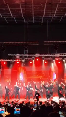 A group of performers in black outfits with red sashes are on a stage performing a synchronized martial arts or dance routine. They are illuminated by bright red stage lights and a large screen behind them displays a glowing, abstract graphic. The stage is set up with lighting rigs and trusses above. In the foreground, an audience is visible, some holding up phones and cameras to record the performance. The ceiling appears to be a large grid of square panels, possibly solar panels.