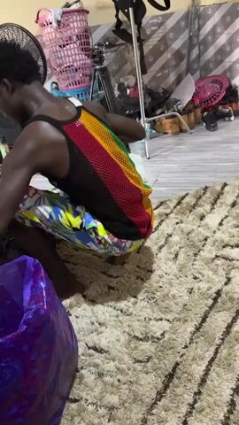 A person kneels on a beige shag rug, wearing a Rasta-colored mesh tank top, and a pair of floral print pants. Behind the person are assorted items, including a black camera tripod, blue plastic laundry baskets, tan boots, and various other film or photography-related items. The background also shows a patterned wall. Another person, wearing khaki pants, walks behind the kneeling person in the middle of the frame.
