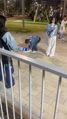The video captures an evening scene in an outdoor park or playground, likely in Kiryat Motzkin, Israel. In the foreground, a white metal fence partially obscures the view. Beyond the fence, several children and adults are present on a paved area and a grassy section with playground equipment in the background.

Two children are prominently featured, playing with blue and white spray foam cans, spraying the foam on each other and the ground. White foam residue is visible on the paved surface, suggesting a playful foam fight. One child wears a denim jacket and white pants, holding a spray can, while another child in a denim outfit actively sprays foam.

In the bottom left, a young child with curly hair, wearing a light green top, is seen near the white fence, being held by an adult's hand on a yellow textured surface. This child appears to be observing the activity.

In the background, other people are seen milling around. One person has a black backpack with the Hebrew word "אגוז" (Egoz) visible on it. The park is illuminated by artificial lights, indicating it is nighttime or evening. The overall atmosphere appears lively and playful.