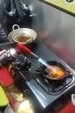 A close-up shot shows a person holding a propane torch with a red handle and a silver canister. The torch is pointed at a small, black frying pan on a black gas stovetop. The pan contains a bubbling, brown liquid and a piece of food that is being seared by the torch's blue and orange flame. To the left of the stovetop, there is a silver pot with a wire strainer inside, also containing a brown liquid. The background is blurry, but appears to be a kitchen or food preparation area, with stainless steel siding visible on the right. The overall scene suggests food is being cooked or flambéed.