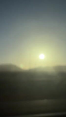 The video shows a sunrise or sunset scene with the sun prominently displayed in the sky. The foreground is blurred, obscuring any specific details of the landscape or objects. The sky is a gradient of blue and yellow, suggesting the sun is low on the horizon. There are lens flares visible, indicating the camera was pointed directly at the sun. The overall impression is of a vast, open environment, possibly a desert or open plain, with a sense of distance and atmospheric haze.