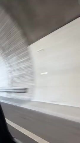 This video captures a scenic drive through Kongsberg, Norway. The footage begins inside a vehicle, looking out the passenger window as it travels through a tunnel with a bright, white interior. The blur of motion suggests the vehicle is moving at a good speed.

Upon exiting the tunnel, the scene opens up to a bright, sunny day. The vehicle is on a paved road with a white line marking the lane. A metal guardrail runs along the side of the road, separating it from a steep, rocky incline covered in vegetation. There are signs visible, one of which is a purple sign with yellow text indicating directions to "Saggrenda" and "Sølvgruvene," suggesting a route to a mining area, likely referencing the historic silver mines of Kongsberg.

The landscape transitions to rolling hills covered in dense green forests under a clear blue sky with scattered white clouds. The sun is shining, casting a warm glow over the scenery, indicating it is likely late afternoon or early evening due to the long shadows and the quality of light. The overall mood is peaceful and picturesque, highlighting the natural beauty of the Norwegian countryside. The drive appears to be a leisurely exploration of the area.