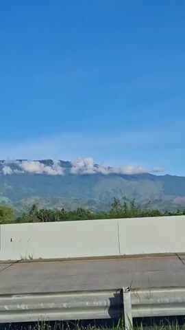 This media file captures a scenic view from a moving vehicle, likely a car or bus, on a highway. The main subjects are a majestic mountain range in the distance, partially shrouded by fluffy white clouds along its peak. The mountain slopes are covered in lush green vegetation, with a dense line of trees in the mid-ground.

In the foreground, a white concrete barrier separates the road from the landscape. The road itself is visible, with clear lane markings, indicating a well-maintained highway. The time of day appears to be morning or early afternoon, given the bright blue sky and the clear, sharp lighting on the landscape. The weather is sunny and pleasant, with a few clouds adding visual interest to the sky.

The setting is a rural or semi-rural area in Leupungbaleu, Indonesia, characterized by its natural beauty and expansive vistas. There are no people or specific objects other than the natural elements and the road infrastructure. The overall mood is one of tranquility and appreciation for nature's grandeur, as one travels through this picturesque location. There is no visible text in the media file.