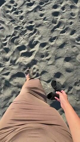 A person wearing a long brown skirt is walking barefoot on a beach. They are holding a pair of black sandals in their right hand. The sand is dark gray and there are footprints and small pieces of debris scattered across it. The footage is taken from a first-person perspective, looking down at the person's legs and feet. There is a wind noise in the background.