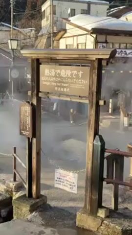 The video shows a hot spring area in Japan. A wooden sign in both Japanese and English warns of the danger of very hot water. Steam rises from the pools behind the sign, partially obscuring the view of buildings and people in the distance. The area is fenced off with wooden railings and a chain. A clock and another sign are visible to the left of the main warning sign. The scene is bathed in sunlight.