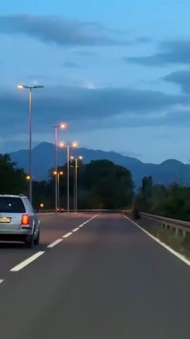 The camera faces the back of a silver station wagon driving on a paved road that curves gently to the right, where the speed limit is 50. The car has its brake lights on. The road is flanked by evenly spaced street lights. The background reveals a dense line of dark green trees with a distant mountain range under a blue, cloudy dusk sky. The scene is still and quiet.